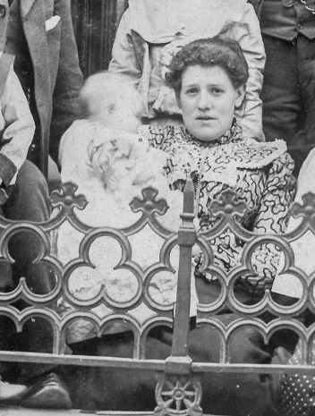 Sarah in a high-necked dress, seated behind a decorative iron railing and holding baby Dora  in her arms.