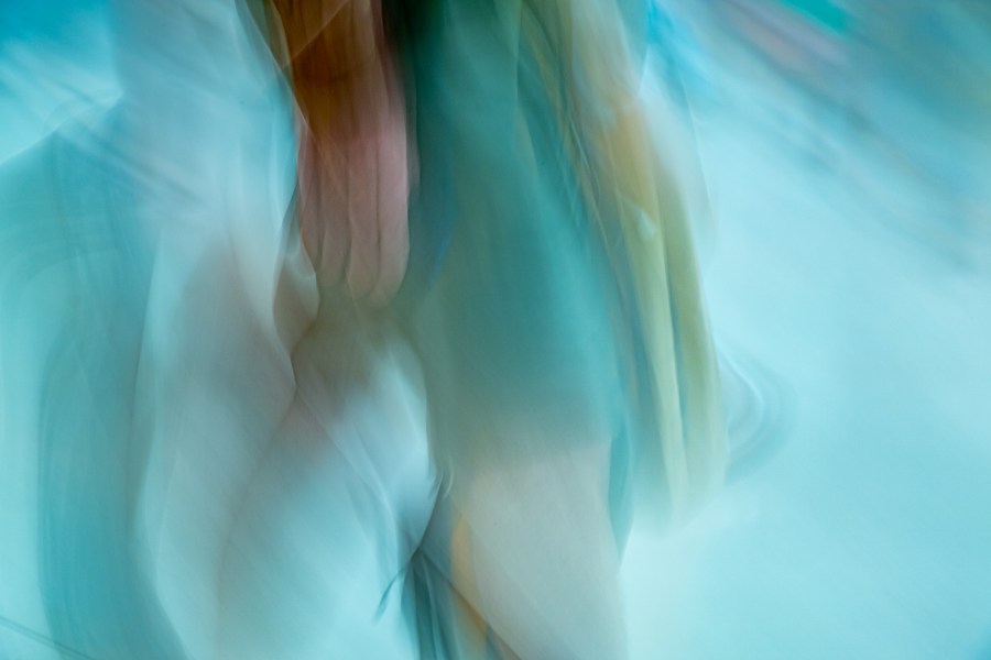A blurred impression of a woman walking by, just the lower torso in a turquoise dress, and calves, with arm and hand nearest the camera in motion too. The bluey-green pale tones predominate, with just the flesh tones varying.