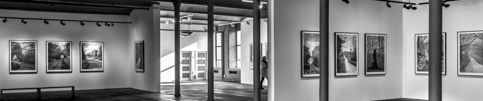 A black and white image of a Hockney Gallery in a former textile mill. Hockney images line the walls, punctuated by cast iron pillars.